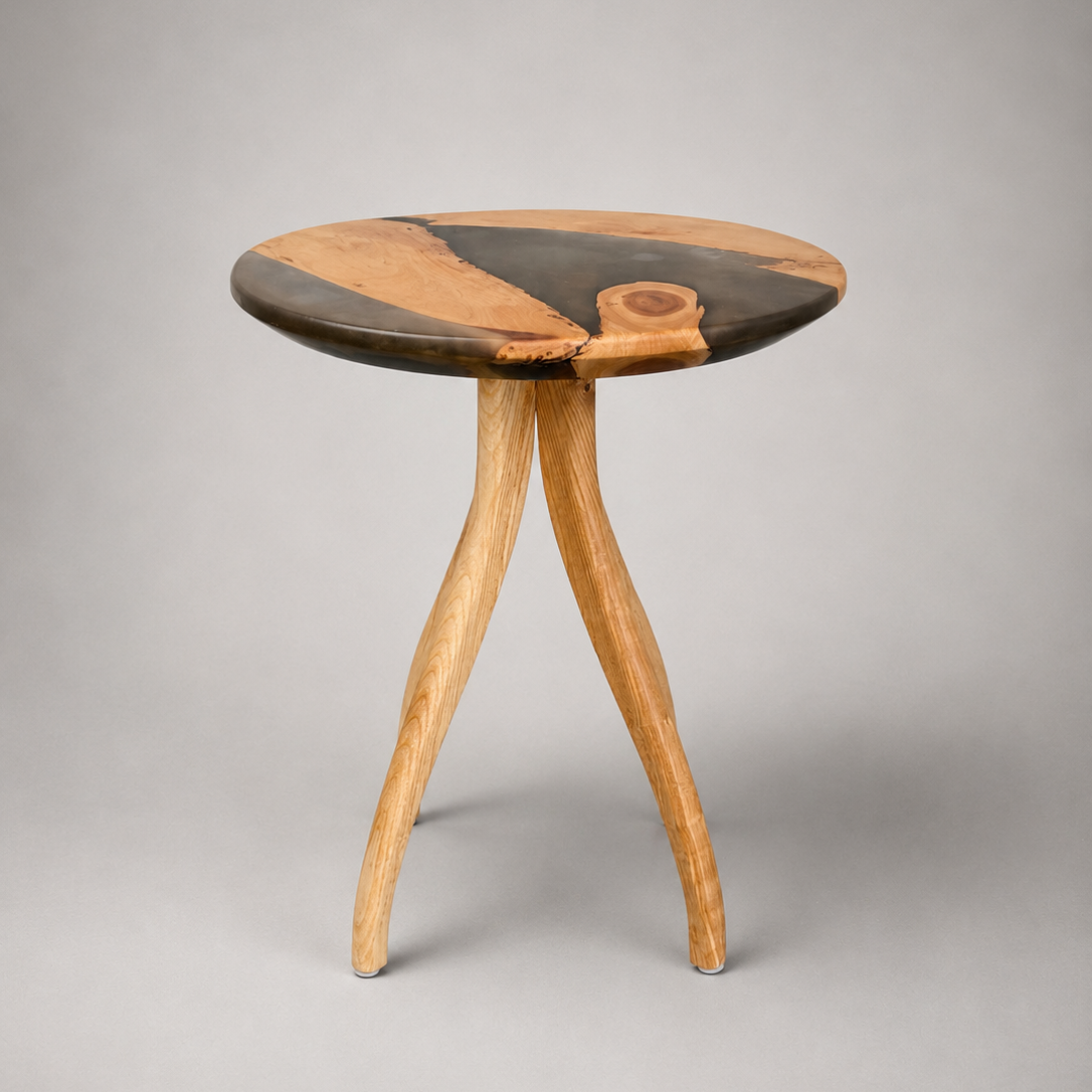 A small round wooden side table with three curved legs, featuring a dark and light wood grain pattern on the tabletop, photographed against a neutral gray background.