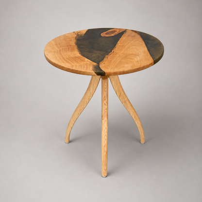 A round wooden side table with a natural live-edge top featuring light and dark wood sections, supported by three splayed legs, photographed on a neutral gray background.