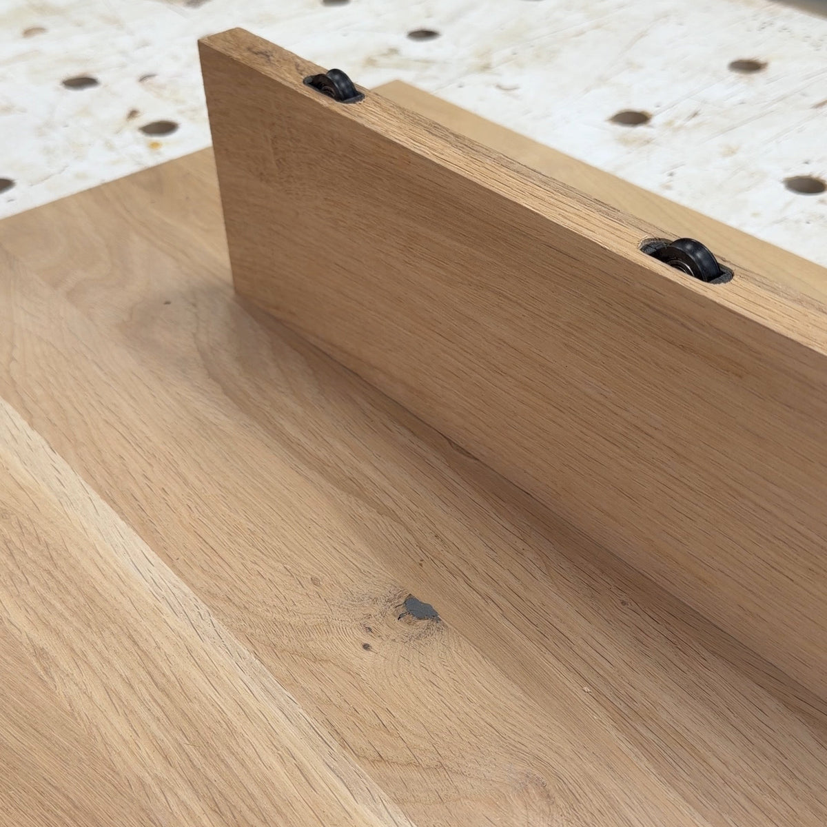 A close-up of a light natural wood board or tabletop with a raised edge and two visible black fasteners, photographed in a workshop setting with a workbench and shelving in the background.