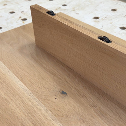 A close-up of a light natural wood board or tabletop with a raised edge and two visible black fasteners, photographed in a workshop setting with a workbench and shelving in the background.