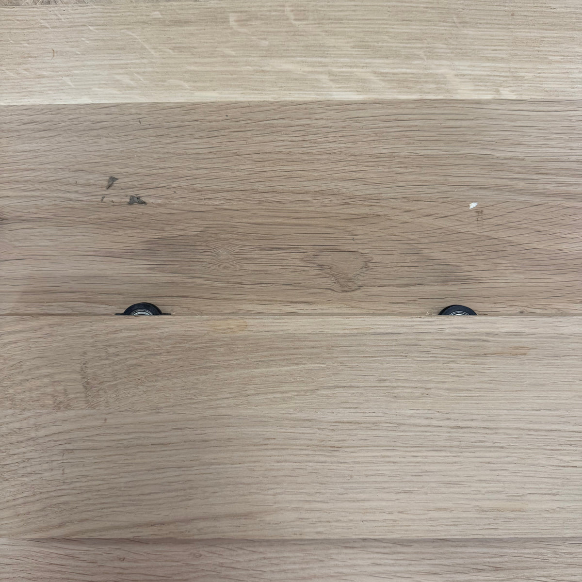 Close-up of a light oak wood surface with visible wood grain, plank seams, and two small dark circular hardware pieces near the lower middle.