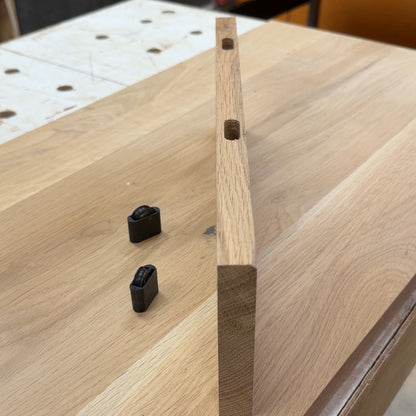 A light natural wood rectangular block with two pre-drilled holes, standing upright on a workbench next to two small black plastic pieces, with a workshop background.